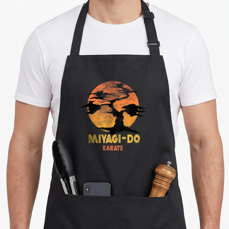 Tablier de cuisine noir avec logo Miyagi-Do Karate sur fond de soleil orange et silhouettes d'arbres stylisés.