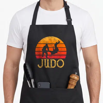 Tablier de cuisine noir avec illustration vintage Judo montrant deux silhouettes en action sur fond de soleil orange et rouge.