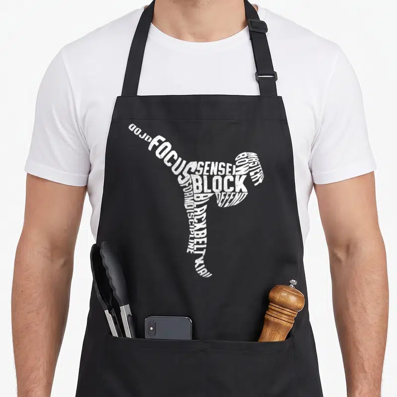 Tablier de cuisine noir avec une silhouette de coup de pied constituée de mots blancs tels que Focus, Block et Sensei.