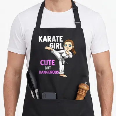 Tablier de cuisine noir avec une illustration de Karate Girl en tenue blanche et ceinture noire effectuant un coup de pied élevé.