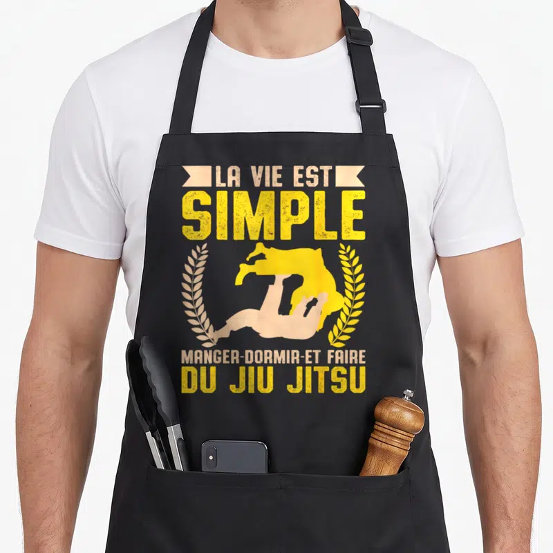 Tablier de cuisine noir avec silhouette jaune de jiu-jitsu et texte La Vie Est Simple Manger Dormir Du Jiu Jitsu encadré de lauriers.