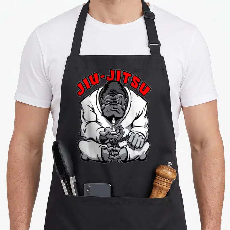 Tablier de cuisine noir avec illustration d'un gorille en kimono blanc tenant une pièce d'échecs et le texte JIU-JITSU en rouge.