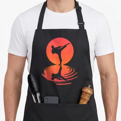 Tablier de cuisine noir avec silhouette d'arts martiaux effectuant un coup de pied face à un disque solaire orange reflété.