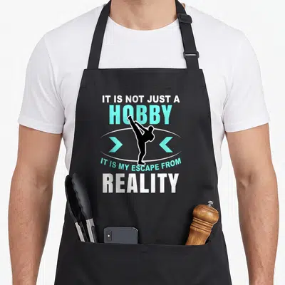 Tablier de cuisine noir avec silhouette de combattant effectuant un coup de pied haut et texte bilingue en blanc et turquoise.