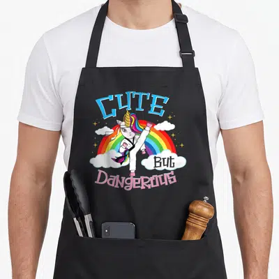 Tablier de cuisine noir avec une licorne effectuant un coup de pied haut devant un arc-en-ciel et le texte « Mignon Mais Dangereux ».