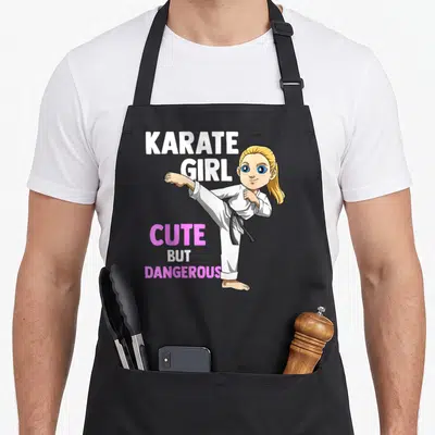 Tablier de cuisine noir avec illustration d'une fille en kimono blanc effectuant un coup de pied et le texte Karate Girl Adroite Mais Dangereuse.