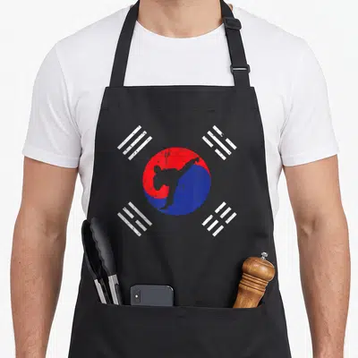 Tablier de cuisine noir avec une silhouette de taekwondo sur un cercle rouge et bleu entouré de symboles blancs.