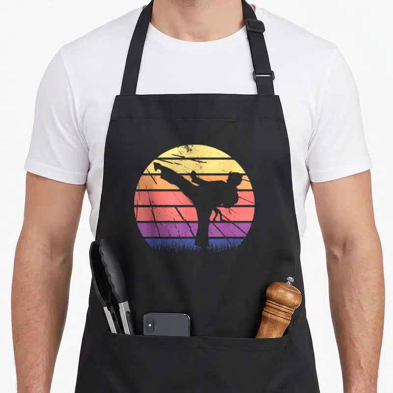 Tablier de cuisine noir avec silhouette de pratiquant d'arts martiaux devant un soleil couchant coloré style vintage.