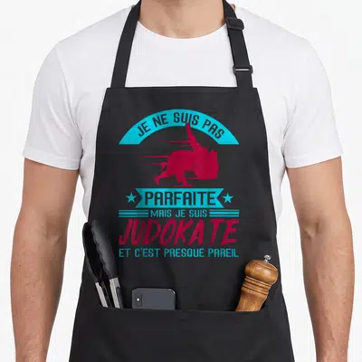 Tablier de cuisine noir avec silhouette judokate rose et texte bleu cyan sur fond uni.