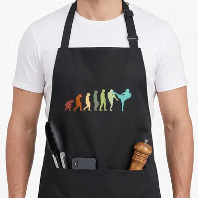 Tablier de cuisine noir avec une illustration colorée montrant l'évolution d'un primate jusqu'à un pratiquant de kickboxer effectuant un coup de pied élevé.