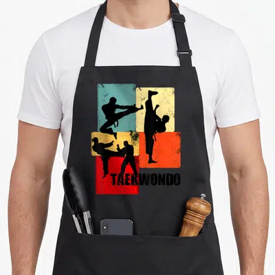 Tablier de cuisine noir avec silhouettes de pratiquants de Taekwondo sur fonds colorés et inscription TAEKWONDO en bas.