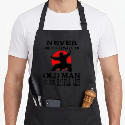 Tablier de cuisine noir avec silhouette martiale rouge et texte anglais humoristique sur fond uni.