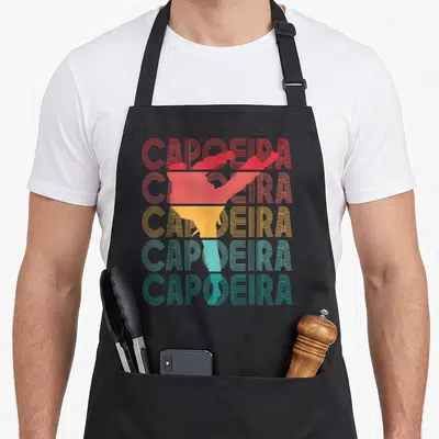 Tablier de cuisine noir avec le mot Capoeira répété en couleurs et une silhouette stylisée au centre.