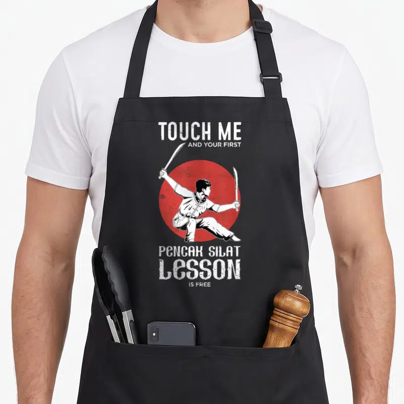 Tablier de cuisine noir avec illustration d'un homme en position de combat tenant deux couteaux devant un cercle rouge.
