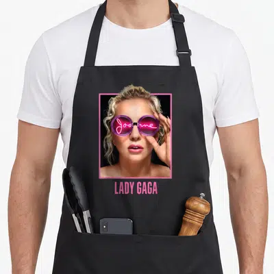 Tablier de cuisine noir avec portrait Lady Gaga lunettes roses inscription Joanne néon et nom LADY GAGA en bas.