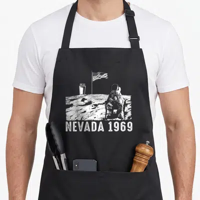 Tablier de cuisine noir avec illustration blanche d'un astronaute assis sur la lune devant un drapeau américain et une enseigne On Air.