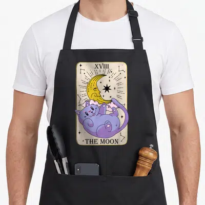 Tablier de cuisine noir avec une illustration de carte de tarot montrant un chat violet sous une lune jaune et l'inscription THE MOON.