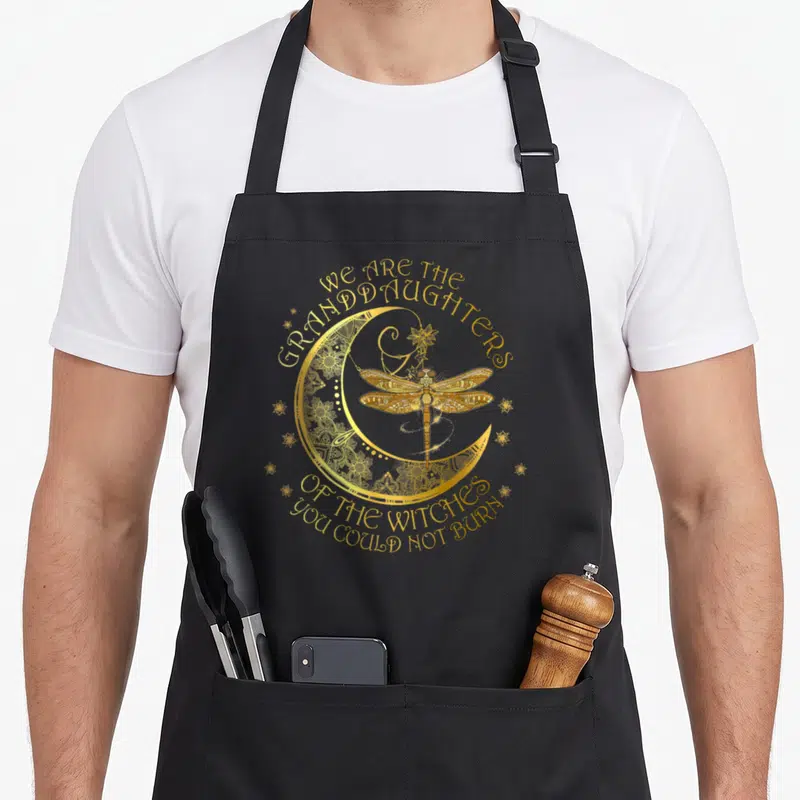 Tablier de cuisine noir avec une libellule dorée, un croissant de lune et un texte ésotérique.