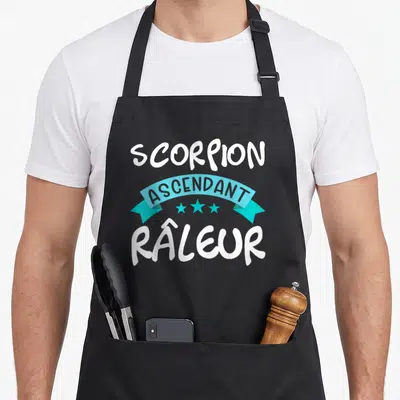 Tablier de cuisine noir avec inscription Scorpion ascendant râleur sur bandeau turquoise et trois étoiles blanches.