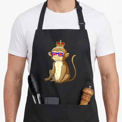 Tablier de cuisine noir avec un singe assis coiffé d'une couronne et portant des lunettes au drapeau britannique.
