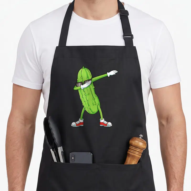 Tablier de cuisine noir avec un concombre vert anthropomorphisé effectuant une pose de dabbing, portant des lunettes noires et des baskets rouges.