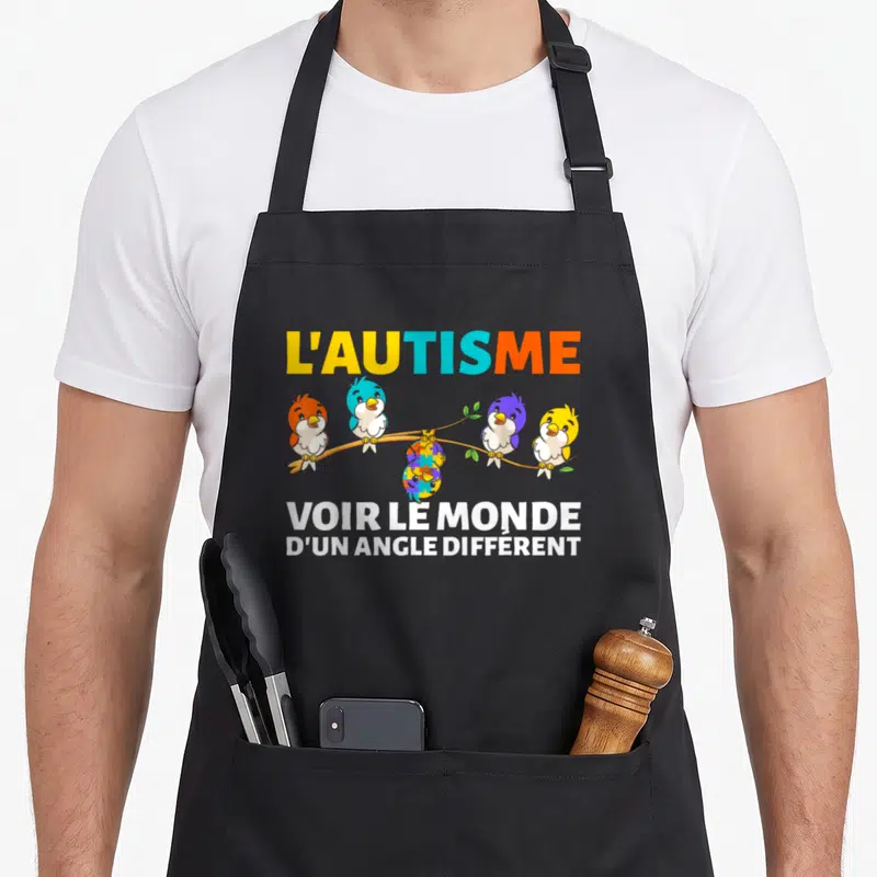 Tablier de cuisine noir avec quatre oiseaux colorés sur une branche et un puzzle central sous le texte Voir le monde d'un angle différent.