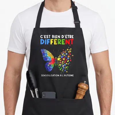 Tablier de cuisine noir avec un papillon multicolore en pièces de puzzle et le texte C'est bien d'être différent.