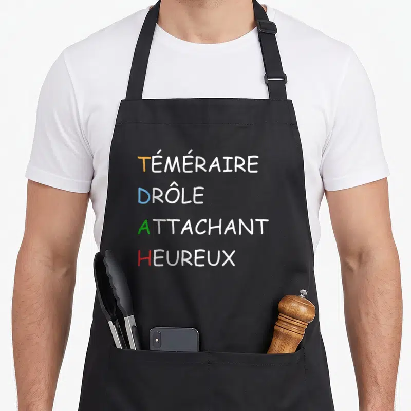 Tablier de cuisine noir avec les mots Téméraire Drôle Attachant et Heureux en lettres colorées.