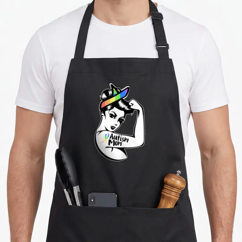 Tablier de cuisine noir avec une illustration de femme aux cheveux chignon portant un bandeau arc-en-ciel et le texte Autism Mom sur le biceps.