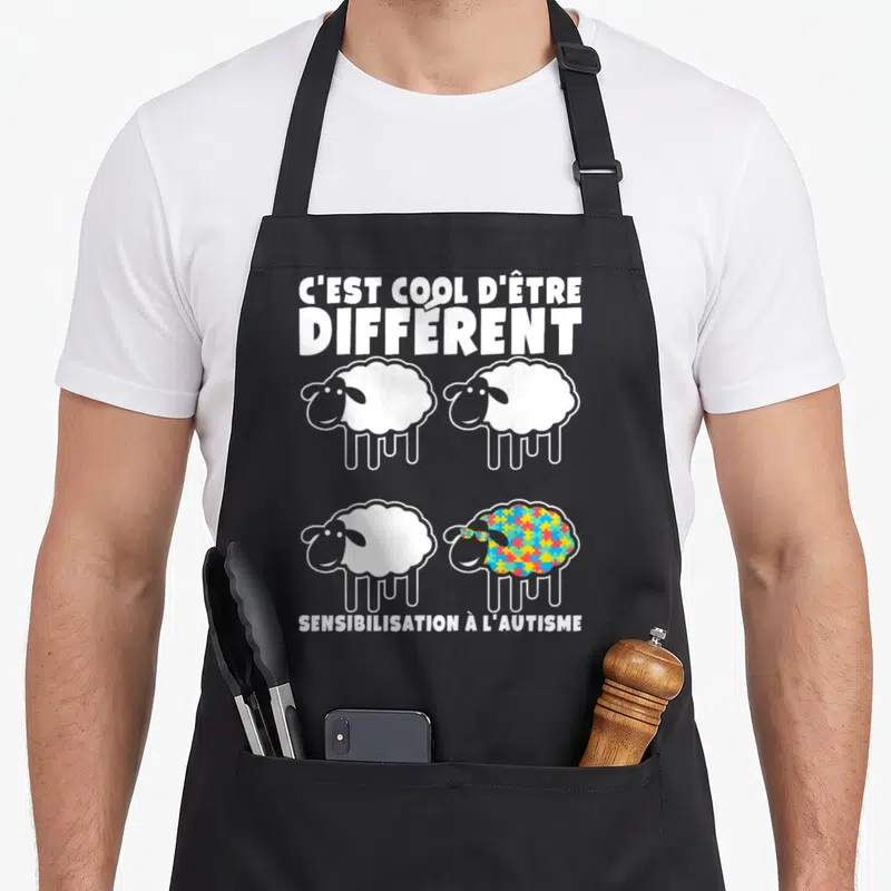 Tablier de cuisine noir avec quatre moutons blancs et un mouton aux pièces de puzzle multicolores sous le texte C'est cool d'être différent.