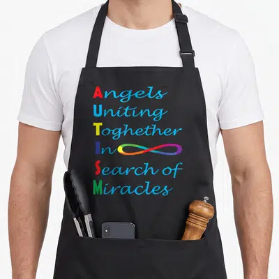 Tablier de cuisine noir avec l'acrostiche ATISM coloré et le message Angels Uniting Together In Search Of Miracles sur fond gris.
