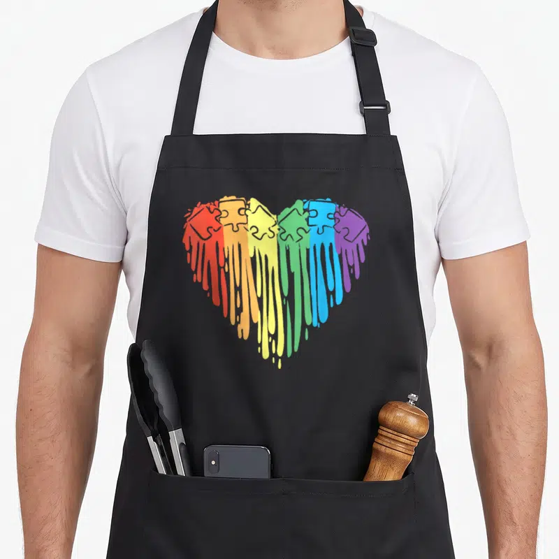 Tablier de cuisine noir avec un cœur arc-en-ciel composé de pièces de puzzle aux couleurs qui s'écoulent vers le bas.