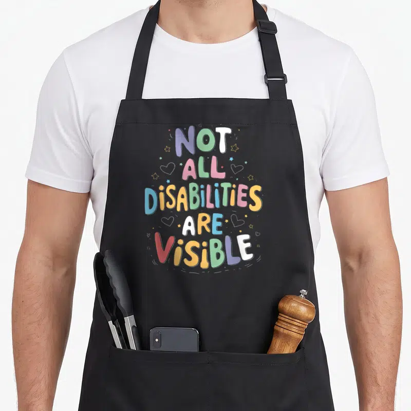 Tablier de cuisine noir avec inscription multicolore Not all disabilities are visible entourée d'étoiles et de cœurs.