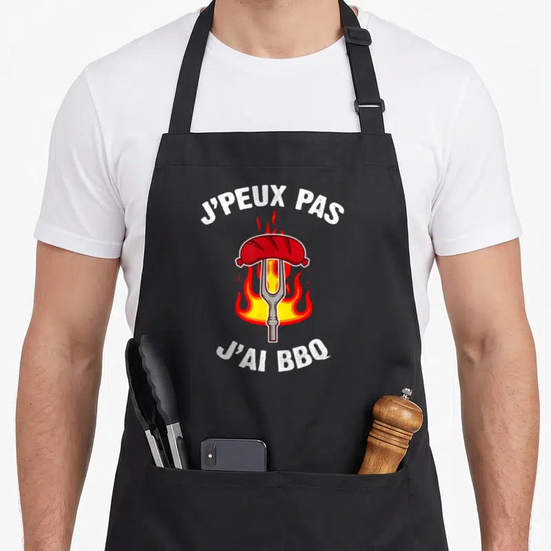 Tablier de cuisine noir avec inscription J'peux pas j'ai BBQ et illustration de saucisse enflammée sur fourchette.