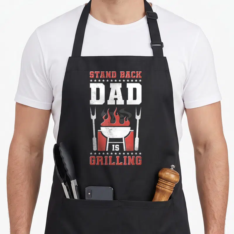 Tablier de cuisine noir avec illustration gril rouge blanc et texte Stand Back Dad Is Grilling encadré d'étoiles.