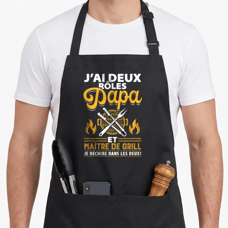 Tablier de cuisine noir avec inscription Papa et maître de grill sur fond blanc et orange, pince et fourche croisées.