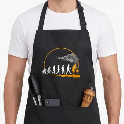 Tablier de cuisine noir avec illustration dorée et blanche représentant l'évolution humaine vers le grillage.
