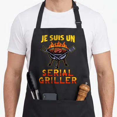 Tablier de cuisine noir avec illustration de grill rond, flamme orange, steak et outils de croisés sous le texte jaune et rouge.