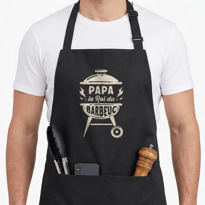 Tablier de cuisine noir avec illustration d'un gril rond beige et texte Papa le Roi du Barbeuc.