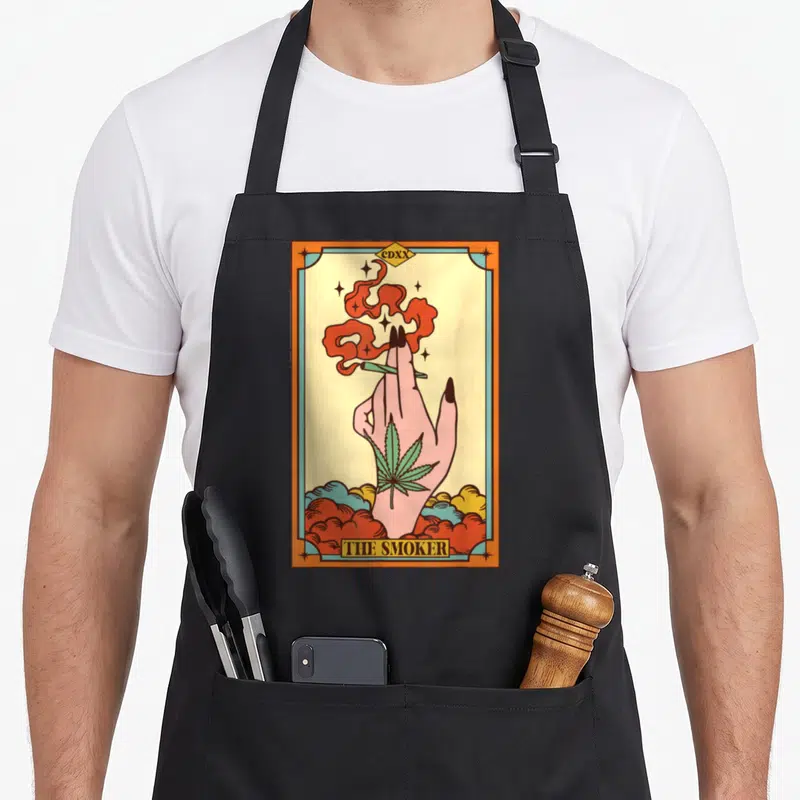 Tablier de cuisine noir avec illustration carte tarot The Smoker main fumant et feuille de cannabis verte.
