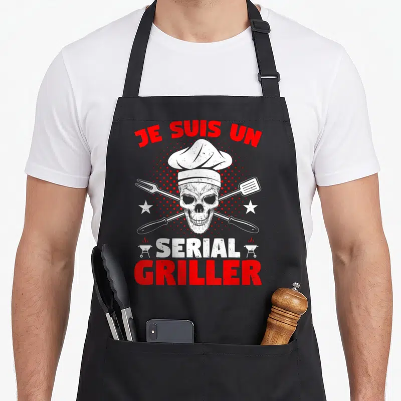 Tablier de cuisine noir avec un crâne en toque et outils de croisés sur fond pointillé rouge.