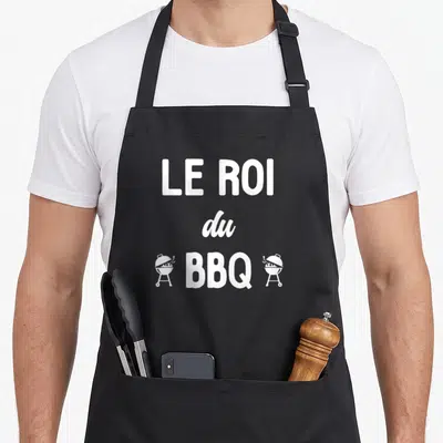 Tablier de cuisine noir avec le texte Le Roi du BBQ encadré par deux icônes de grilles de cuisson blanches.