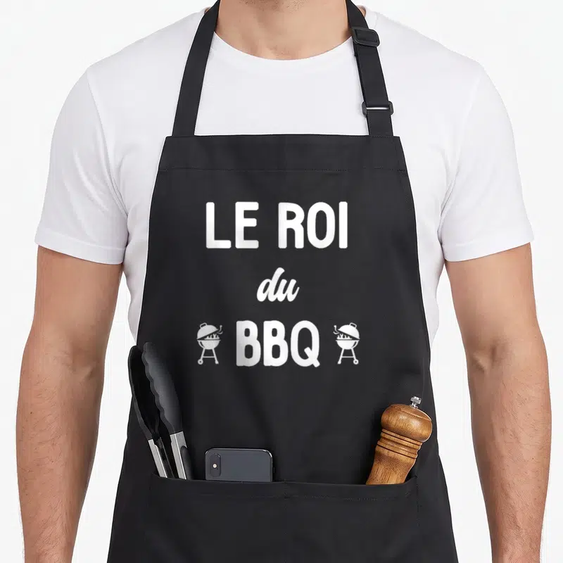 Tablier de cuisine noir avec le texte Le Roi du BBQ encadré par deux icônes de grilles de cuisson blanches.