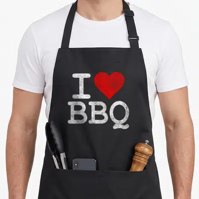 Tablier de cuisine noir avec inscription blanche I Love BBQ et un cœur rouge stylisé au centre du torse.