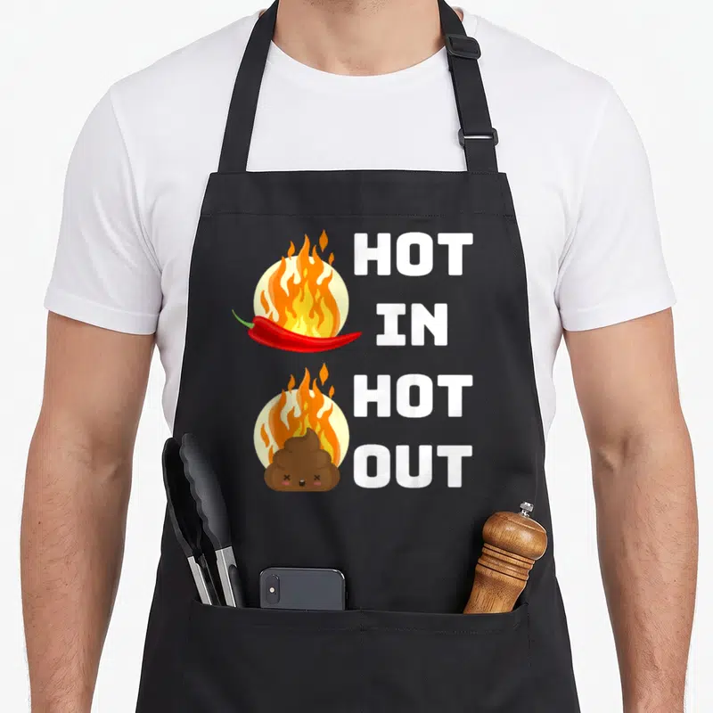 Tablier de cuisine noir avec illustration d'un piment en feu et d'une fiente fumante sous le texte Hot In Hot Out.