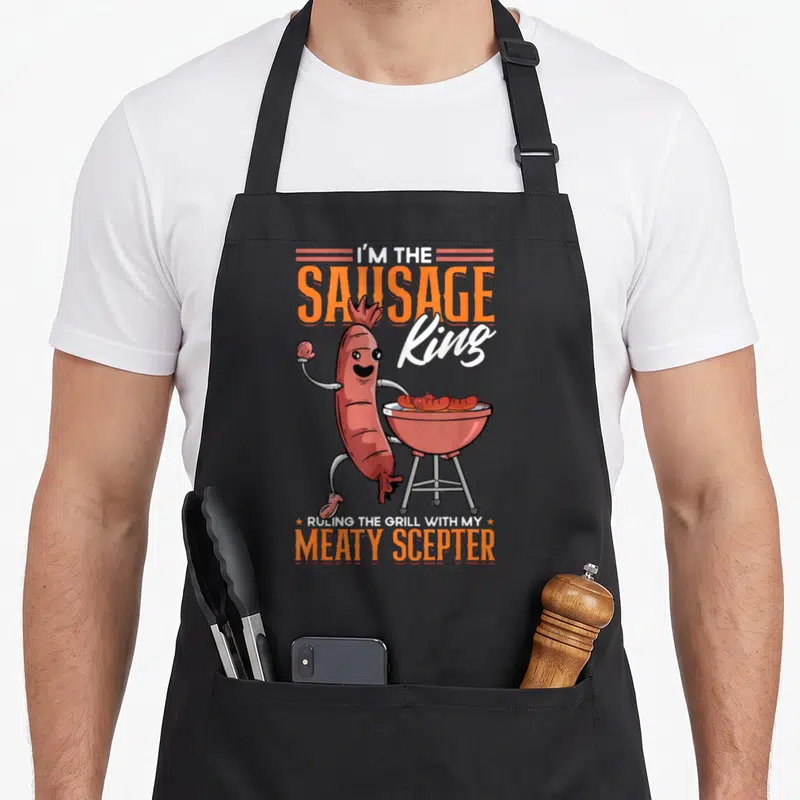 Tablier de cuisine noir avec une saucisse royale debout près d'un gril rouge et des textes orange et blancs.
