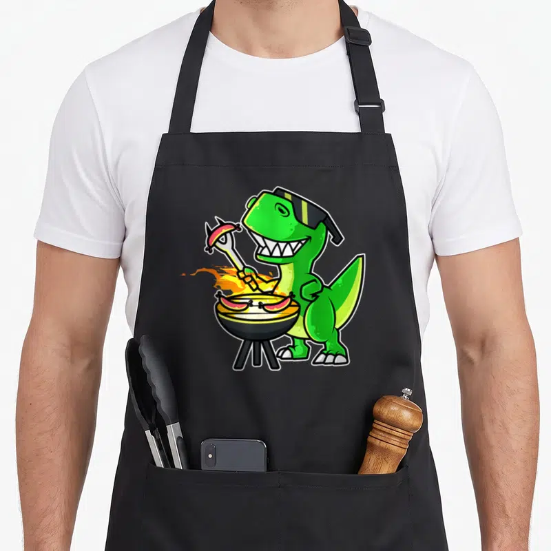 Tablier de cuisine noir avec illustration d'un dinosaure vert grillant des saucisses sur un barbecue rond.