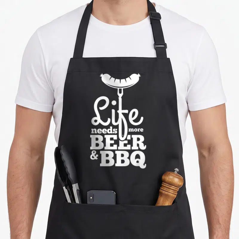 Tablier de cuisine noir avec inscription blanche Life needs more Beer & BBQ et saucisse sur fourchette stylisée.