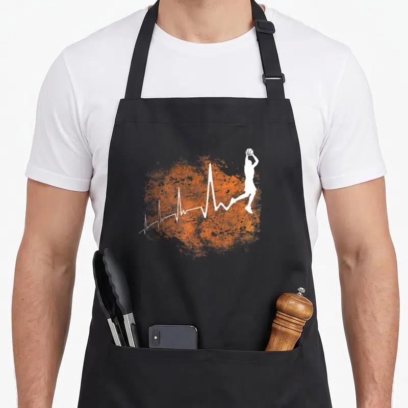 Tablier de cuisine noir avec silhouette blanche d'un joueur de basket-ball en saut et ligne de battement de cœur sur fond orange texturé.