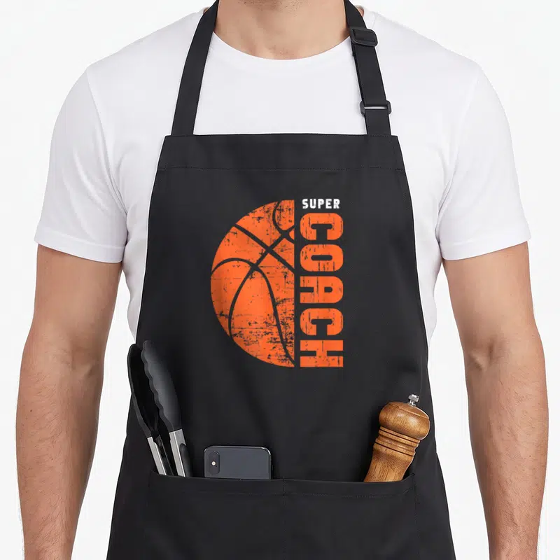 Tablier de cuisine noir avec illustration d'un ballon de basket orange et le texte Super Coach en typographie usée.